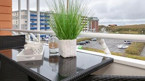 A balcony with a wicker chair, a glass table, a potted plant, and a view of a parking lot and buildings. - Sommerset - 2-room apartment Sommerset, directly on the beach with balcony & parking space (Sylt)