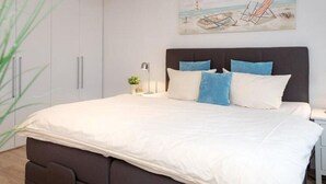 A bedroom with a large bed, a nightstand, a lamp, and a painting on the wall. - Sommerset - 2-room apartment Sommerset, directly on the beach with balcony & parking space (Sylt)