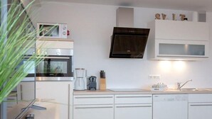 A modern kitchen with white cabinetry, a built-in oven, a microwave, and a stainless steel range hood. - Sommerset - 2-room apartment Sommerset, directly on the beach with balcony & parking space (Sylt)