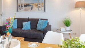 A modern living room with a dark grey sofa, a small round table with a plant, and a painting of a beach scene on the wall. - Sommerset - 2-room apartment Sommerset, directly on the beach with balcony & parking space (Sylt)