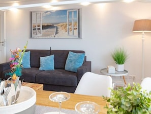 Ein modernes Wohnzimmer mit einem dunkelgrauen Sofa, einem kleinen runden Tisch mit einer Pflanze und einem Gemälde mit einer Strandansicht an der Wand.