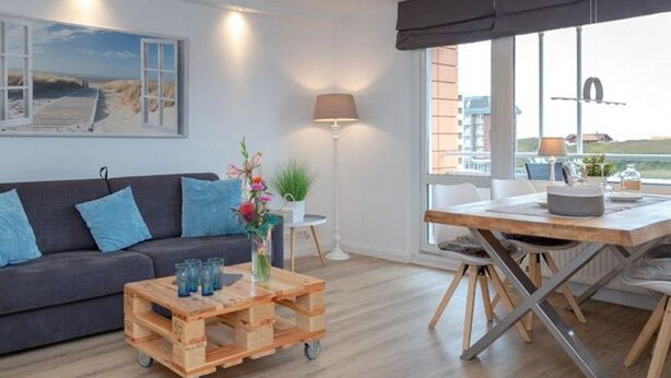 A modern living room with a dark grey sofa, a wooden coffee table, and a dining table with chairs. There is a lamp, a potted plant, and a painting on the wall. - Sommerset - 2-room apartment Sommerset, directly on the beach with balcony & parking space (Sylt)