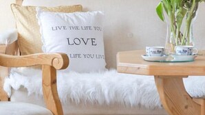 A cozy seating area with a wooden chair, a cushioned bench, and a table with a teapot and cups. A pillow with the text "LIVE THE LIFE YOU LOVE" is visible.