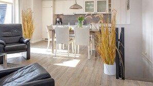 A modern living room with a black leather sofa, a dining area with a white table and chairs, and a kitchen in the background. - Big Wave - BIG WAVE maisonette, central, 2 bedrooms, swimming pool, roof terrace, (Sylt)
