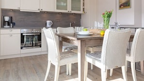 A modern kitchen with white cabinetry, a wooden dining table, wicker chairs, and a hanging pendant light. - Big Wave - BIG WAVE maisonette, central, 2 bedrooms, swimming pool, roof terrace, (Sylt)