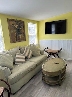 Ein Wohnzimmer mit einer Couch, einem runden Korbstuhl-Tisch, einem an der Wand montierten Fernseher und einem Fenster mit Jalousien.