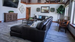 Amplia sala de estar con un sofá de cuero grande, una mesa de centro de madera y un comedor con sillas de madera. Un televisor está montado en la pared y hay una escalera que conduce a un nivel superior.