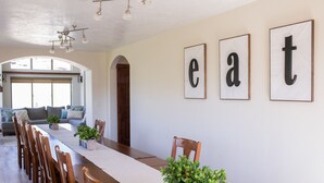Un comedor con una larga mesa de madera, sillas y plantas en macetas. Hay letras enmarcadas en la pared.