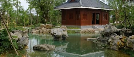 Uma casa de madeira tradicional com telhado de cerâmica, rodeada por um lago com pedras e vegetação.