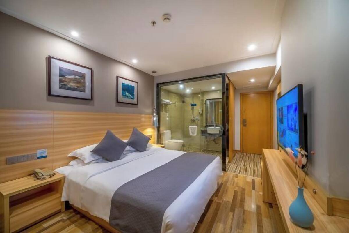 A hotel room with a bed, bedside table, TV, and a bathroom visible through a glass partition.
