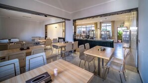 A modern hotel dining area with multiple tables and chairs, a kitchen visible through an open doorway, and a view of the outdoors.