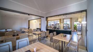 A modern dining area with tables and chairs, a kitchen in the background, and a view of the outdoors.