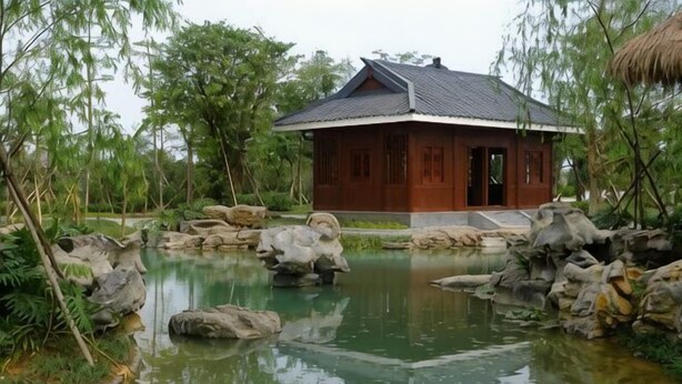 A traditional wooden house with a tiled roof, surrounded by a pond with rocks and greenery.