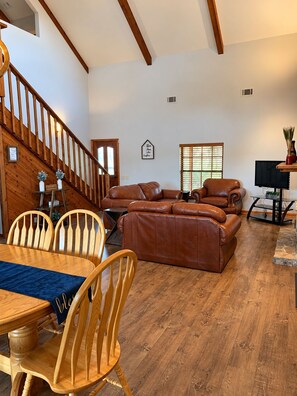A spacious living area with a wooden staircase, leather sofas, a dining table with chairs, and a television.