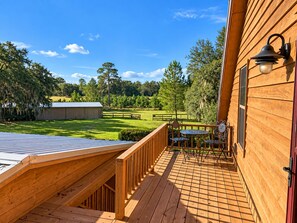 A wooden deck with a staircase leading to a covered area, a table with chairs, and a view of a fenced property with trees and a barn.