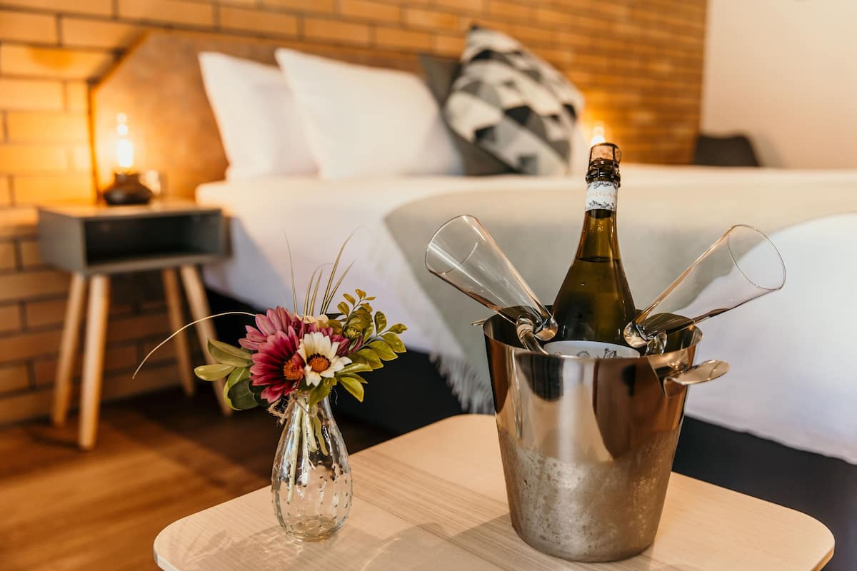 A hotel room with a bed, a bedside table with a bottle of champagne and glasses, and a vase with flowers.