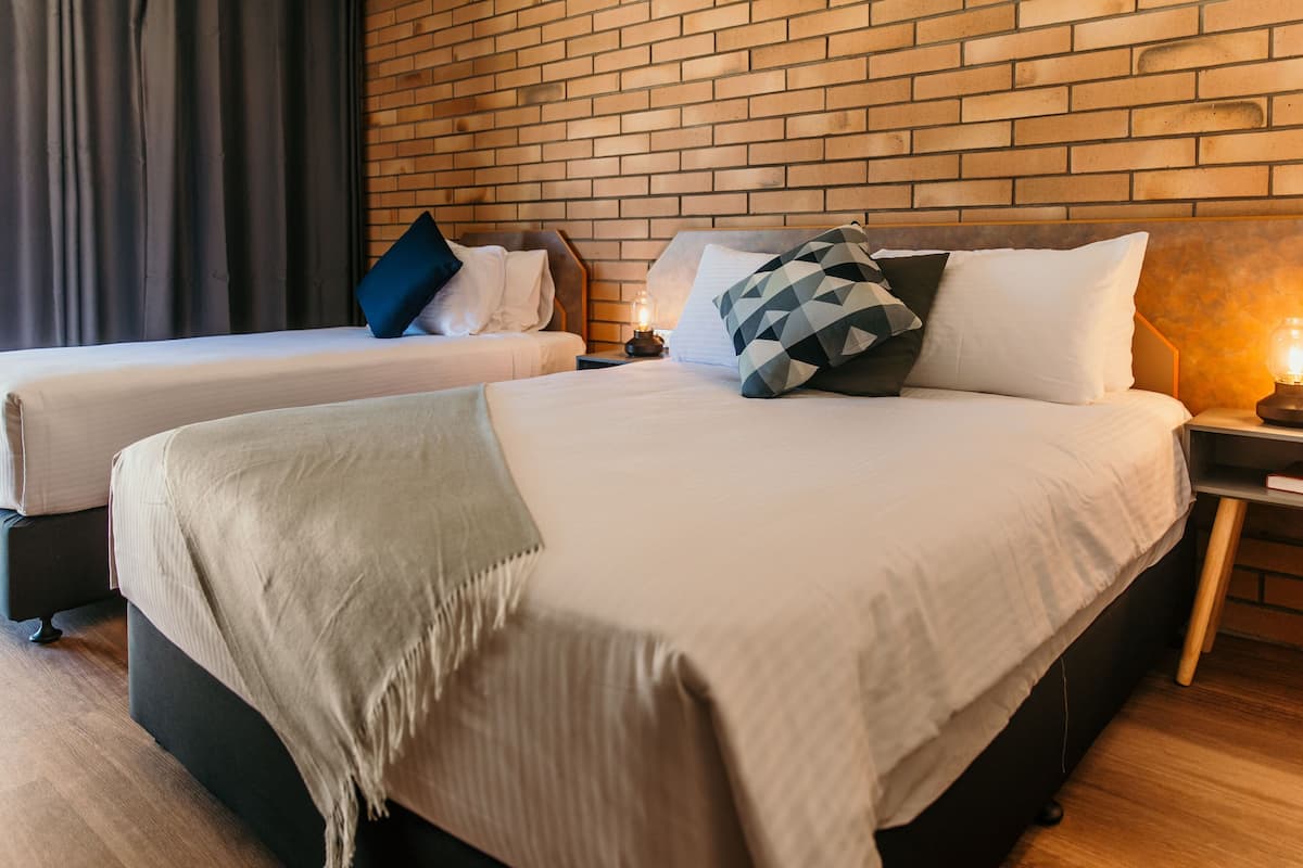 A hotel room with two beds, a brick wall, and a nightstand with a lamp.