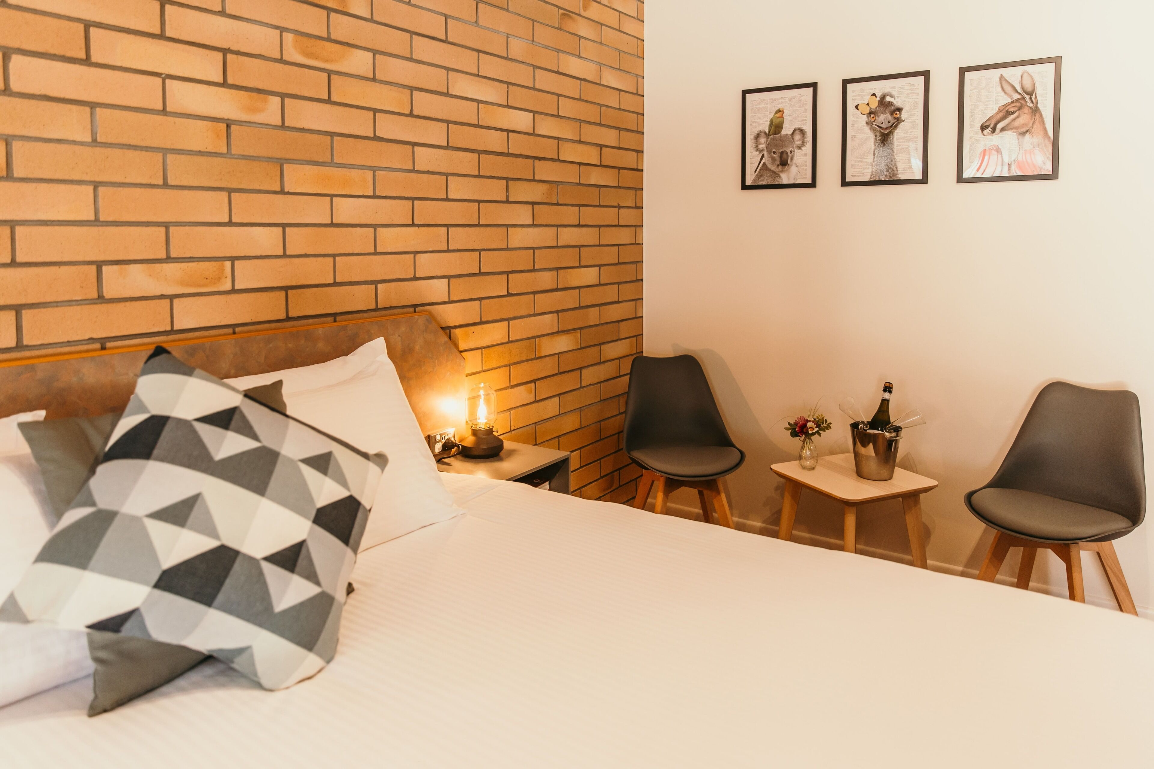 A hotel room with a brick wall, a bed with patterned pillows, a nightstand with a lamp, and two chairs with a small table between them.