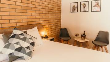 A hotel room with a brick wall, a bed with patterned pillows, a nightstand with a lamp, and two chairs with a small table between them.