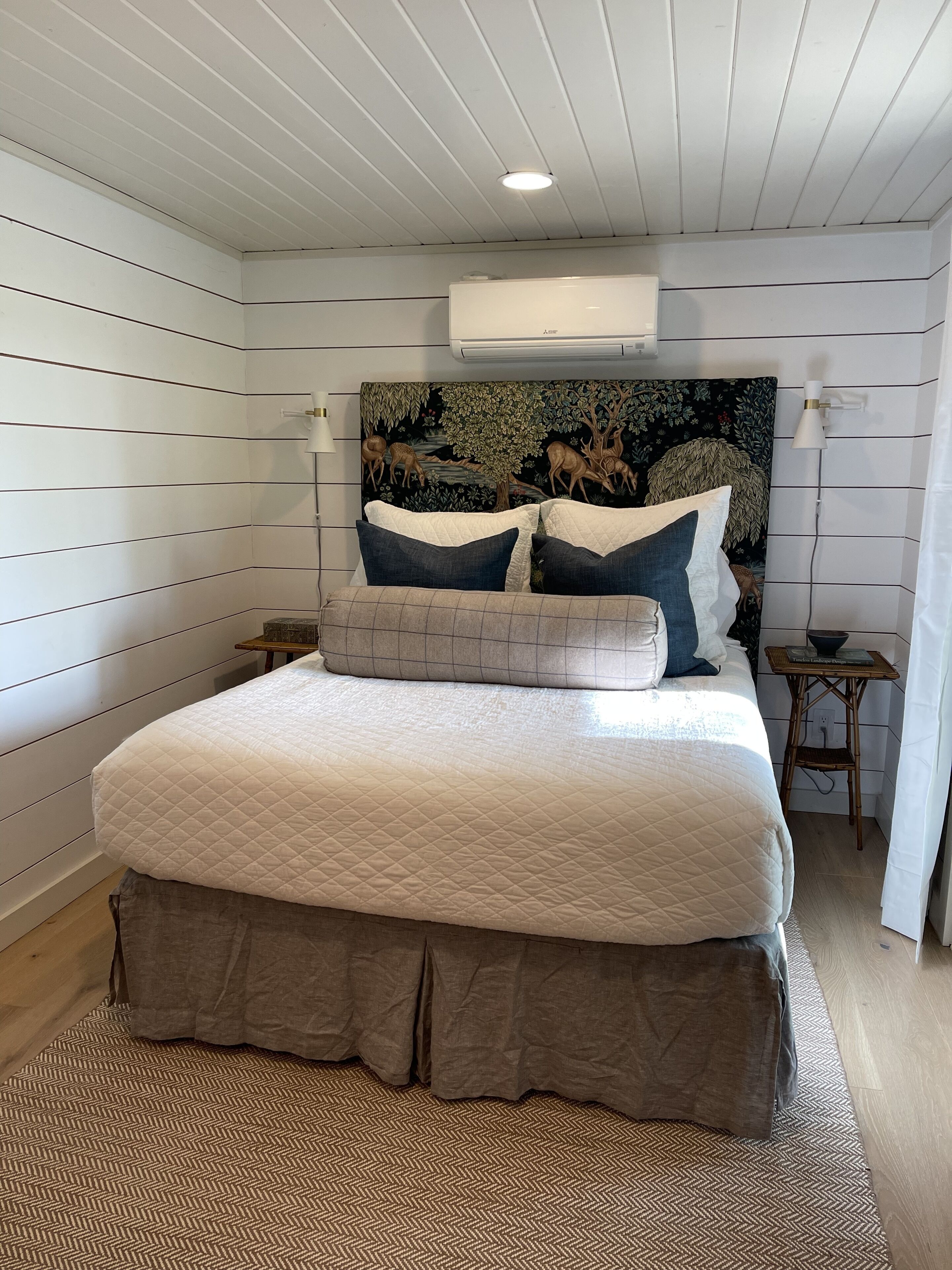 A bedroom with a large bed, a headboard with a deer mural, a wall-mounted air conditioner, and a small table with a lamp.