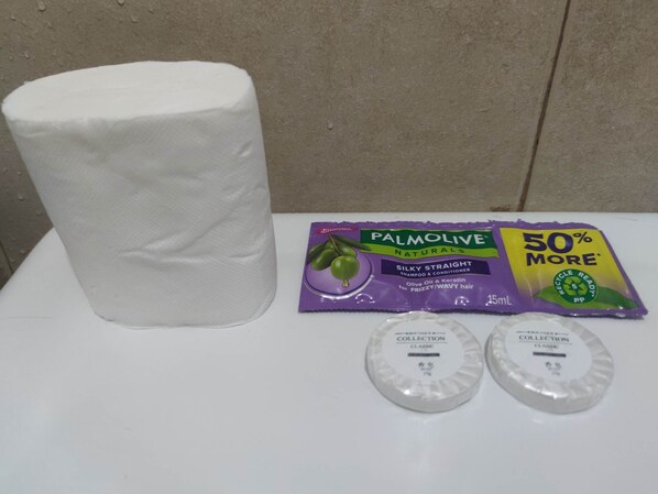 Un baño con un rollo de papel higiénico, un paquete de champú y acondicionador, y dos pequeños recipientes.