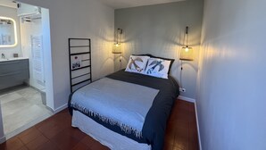 Ein Schlafzimmer mit einem Bett, Nachttischlampen, einem Nachttisch, einem Spiegel und einem im Hintergrund sichtbaren Badezimmer.