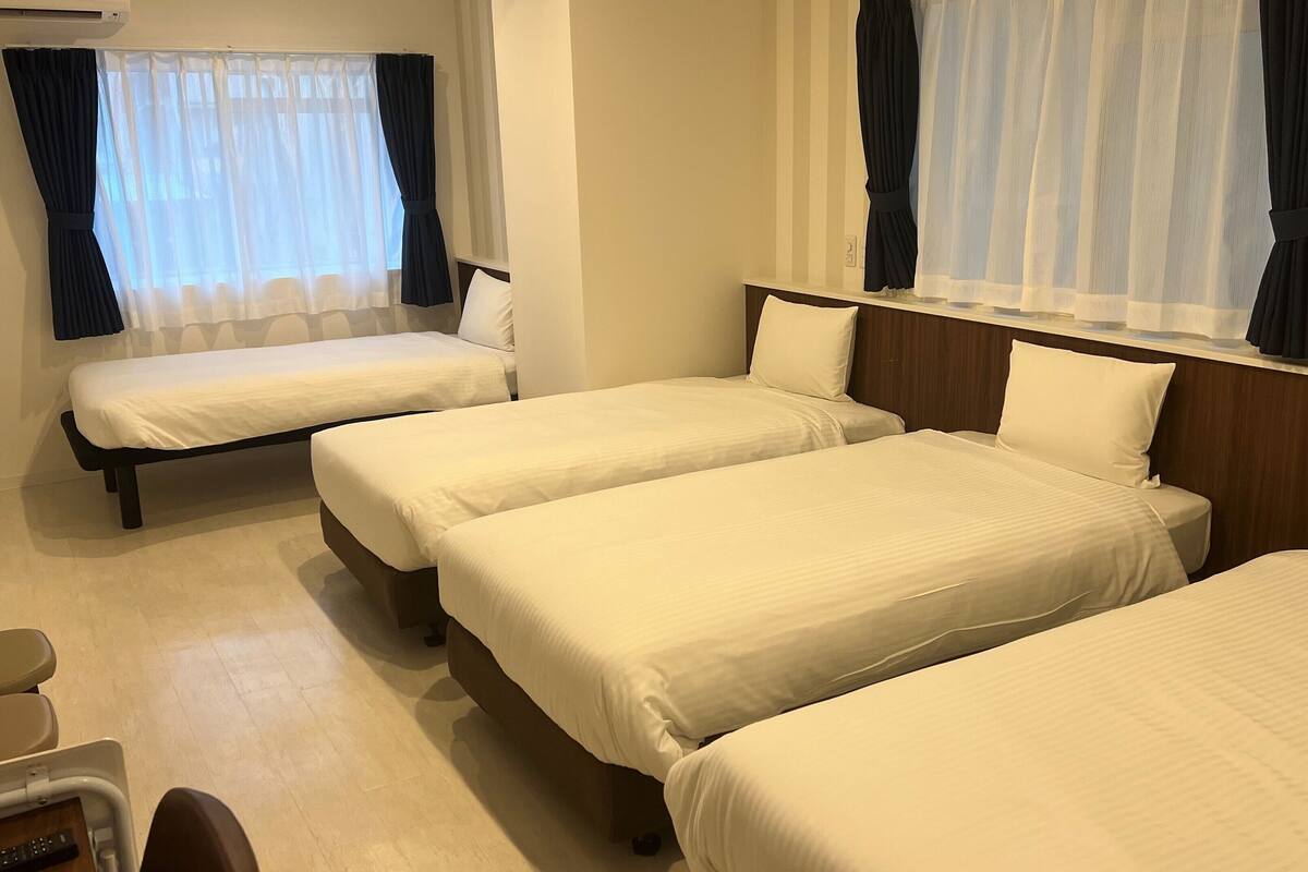 A hotel room with three single beds, a wooden headboard, a small desk with a chair, and a window with sheer curtains.