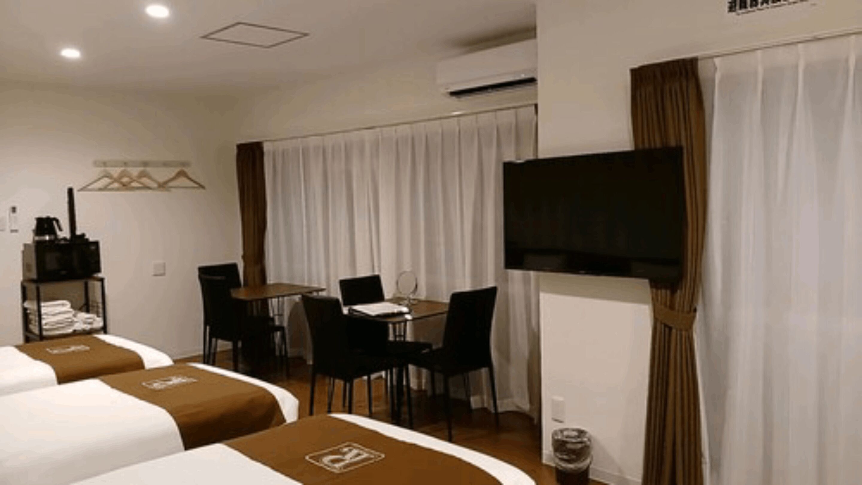 A hotel room with two beds, a dining table with chairs, a TV mounted on the wall, and a window with curtains.