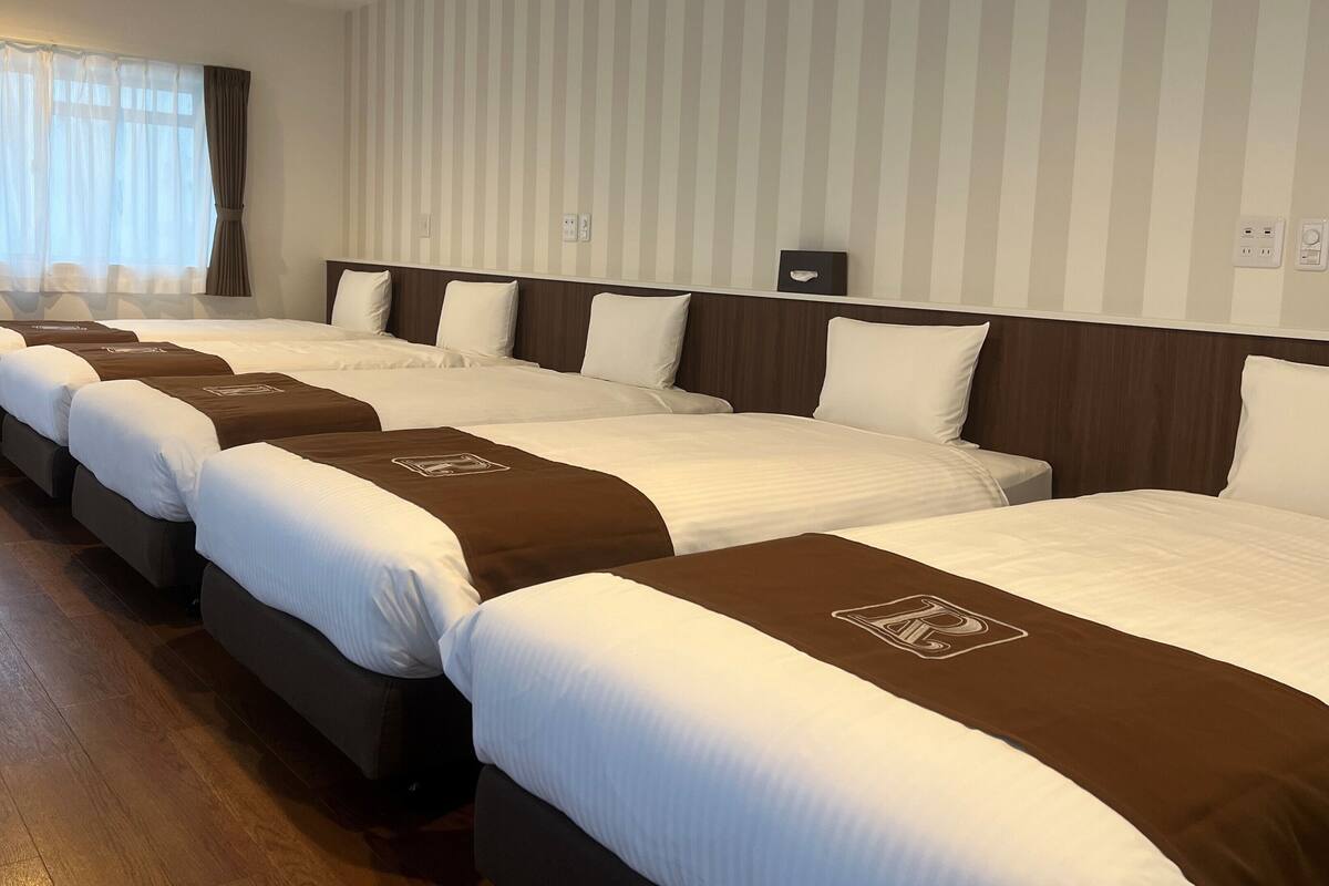 A hotel room with four beds arranged in a row, each with a brown headboard and white bedding.