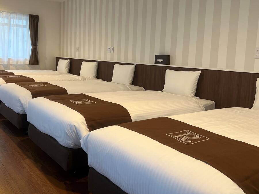Habitación de hotel con cuatro camas dispuestas en fila, cada una con cabecera marrón y ropa de cama blanca.