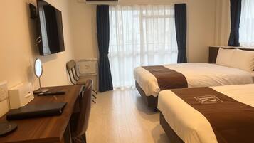A hotel room with two beds, a desk with a chair, a TV, and a window with sheer curtains.