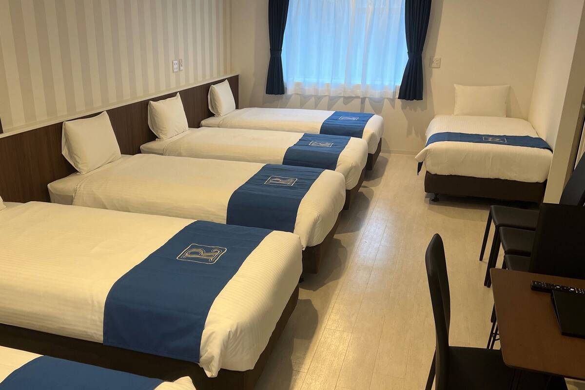 A hotel room with four single beds, each with a blue and white bedspread and matching pillows. The room has a wooden headboard, a small table with a chair, and a window with curtains.