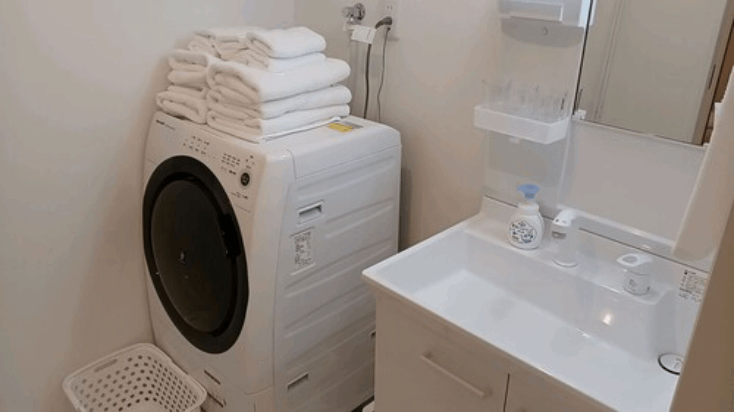 A compact laundry area with a washing machine, stacked towels, a small sink with soap dispenser, and a mirror above the sink.