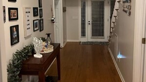 Un pasillo con piso de madera, paredes blancas y una puerta al final. Se observan cuadros enmarcados en la pared y una pequeña mesa con una planta y adornos.