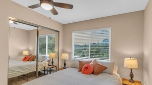 Une chambre à coucher avec un grand lit, deux tables de chevet avec des lampes, un ventilateur de plafond, un grand miroir et une fenêtre avec des stores.