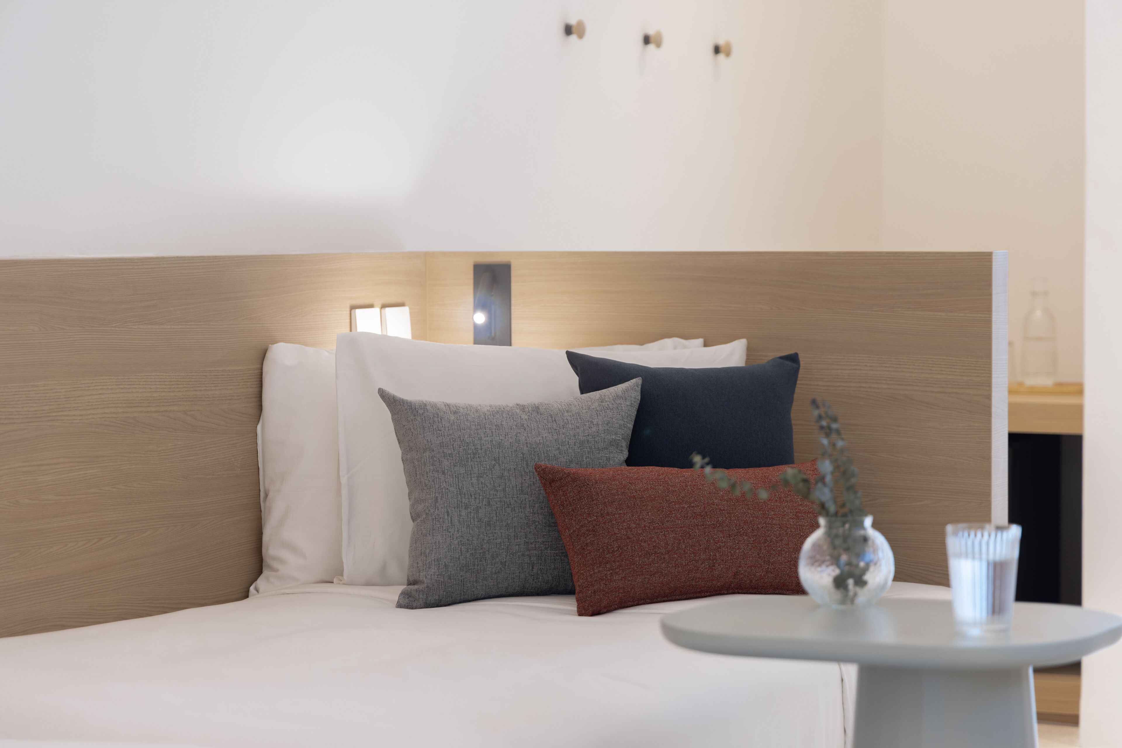 A modern hotel room with a wooden headboard, a white bed, and a small round table with a vase and a glass of water.
