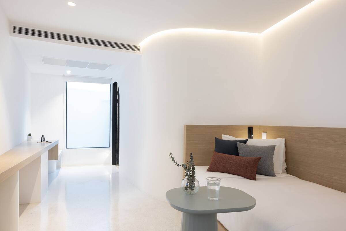 A modern hotel room with a minimalist design, featuring a bed with multiple pillows, a bedside table with a vase and a glass, and a view of a bright hallway.