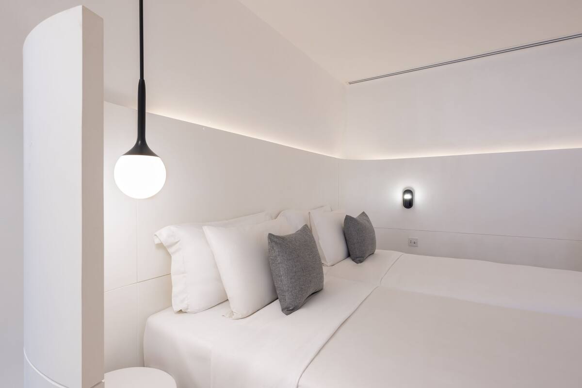 A modern bedroom with a curved ceiling, a bed with white and gray bedding, a round light fixture, and a wall-mounted light.