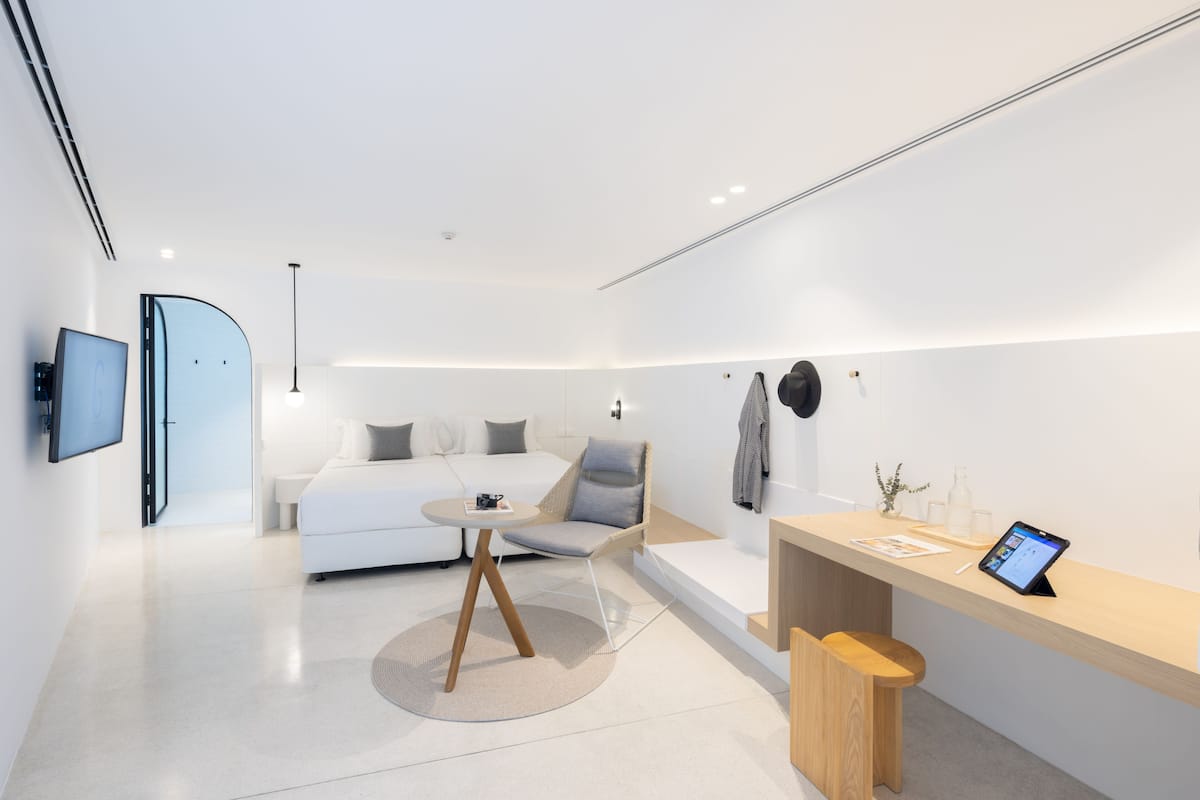 A modern hotel room with a bed, a seating area with a table, a desk with a computer, and a TV mounted on the wall.