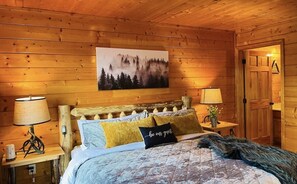 Ein gemütliches Schlafzimmer in einer Holzhütte mit einem Bett, zwei Nachttischlampen, einem Waldgemälde im Bilderrahmen und einer Tür zu einem anderen Raum.