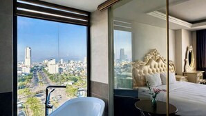 A modern hotel room with a large bathtub, a bed with a tufted headboard, and a city view through a glass door. - Century Hotel (Da Nang)