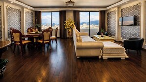 A spacious living room with a large sofa, dining table, and chairs. There is a TV mounted on the wall and a large window offering a city view. - Century Hotel (Da Nang)