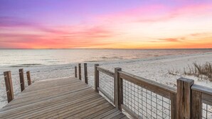 Ein hölzerner Steg führt zu einem Strand, im Hintergrund ist ein Sonnenuntergang zu sehen.