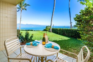 A round table with two chairs, a bottle, and glasses on a patio with a view of the ocean and palm trees.