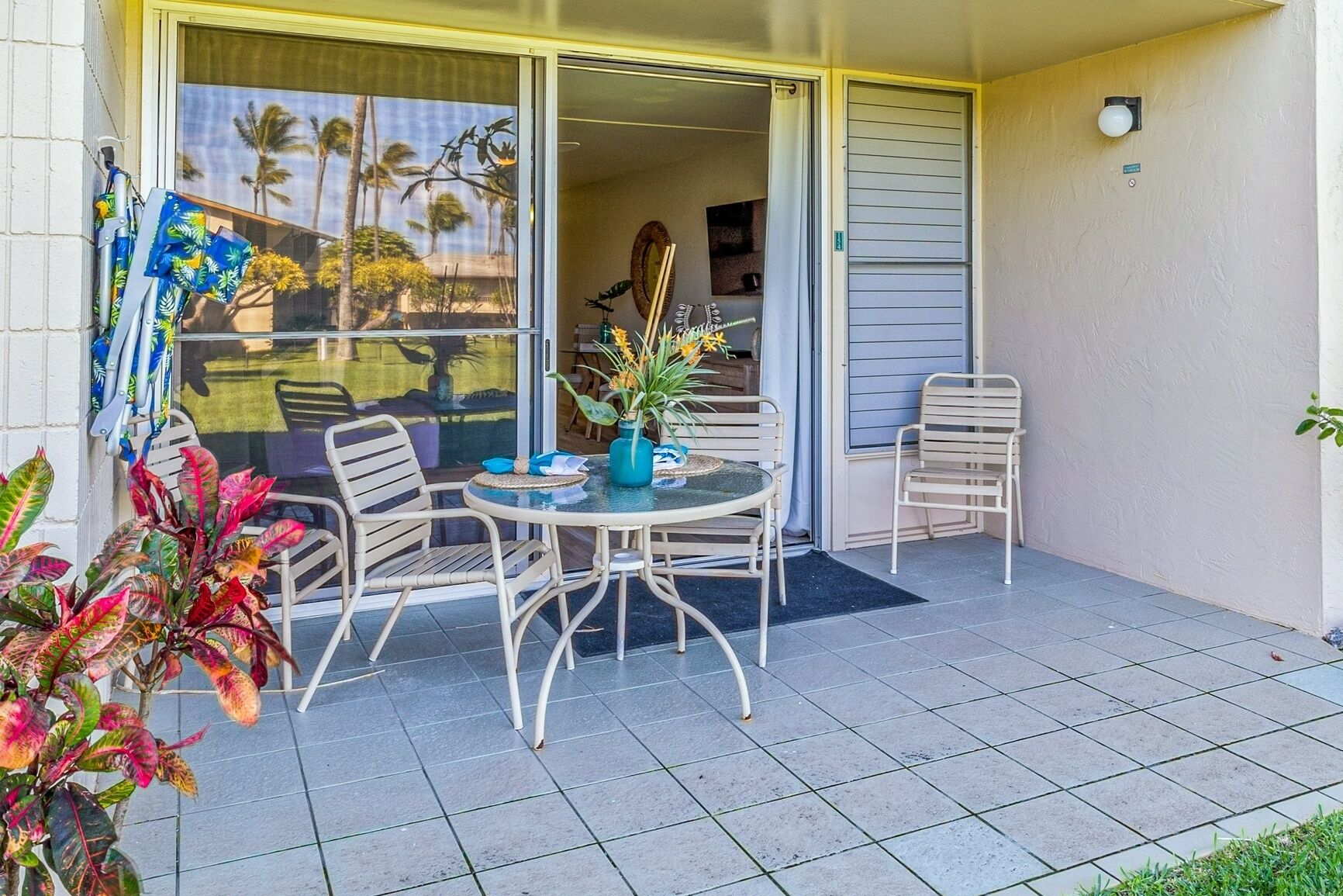 A patio with a round table and four chairs, a sliding glass door, and a view of palm trees.