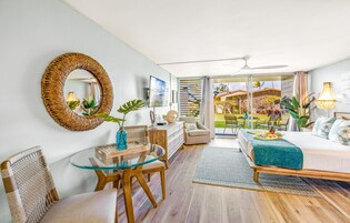 Una habitación de hotel moderna con una mesa de comedor de vidrio, sillas de mimbre, una cama con colcha color aguamarina y un balcón con vistas al jardín.