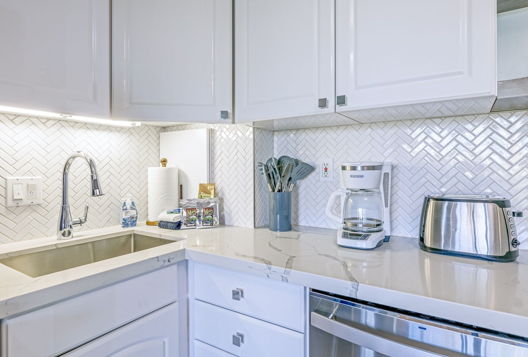 A modern kitchen with white cabinets, a herringbone tile backsplash, a stainless steel coffee maker, and a dishwasher.