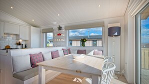A modern kitchen with white cabinetry, a built-in bench with cushions, and a large window offering a view of the outdoors. - Crabtree Lodge -  a lodge that sleeps 6 guests  in 3 bedrooms (Gatehouse Of Fleet)