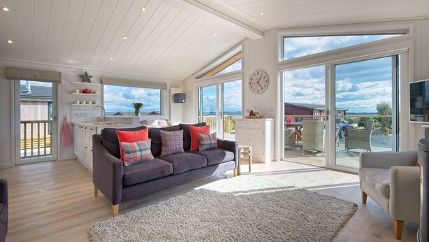 A spacious living area with a grey sofa, a white rug, and a dining area with a table and chairs. - Crabtree Lodge -  a lodge that sleeps 6 guests  in 3 bedrooms (Gatehouse Of Fleet)