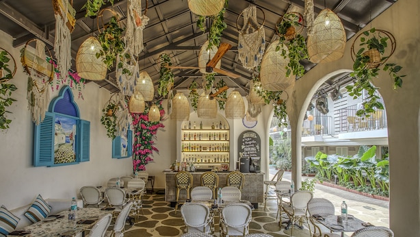 An indoor seating area with wicker furniture, a bar in the background, and hanging plants. - Ocean Palms Goa (Calangute)